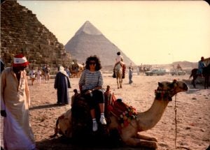 Woman sits on camel in front of pyramids.