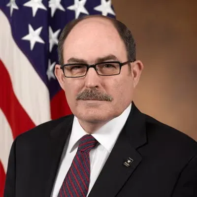 Headshot of David E. Johnson with American flag behind him, wearing black-rimmed glasses, black blazer, white shirt, and maroon tie.
