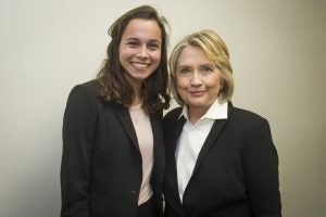 Two women pose for a photo in front of blank wall.