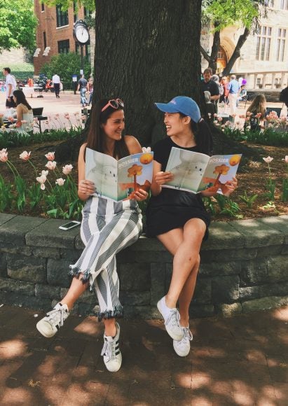 Two women sit reading magazines outside.