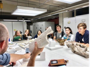 Students sitting around a table watch museum curator present fossil.