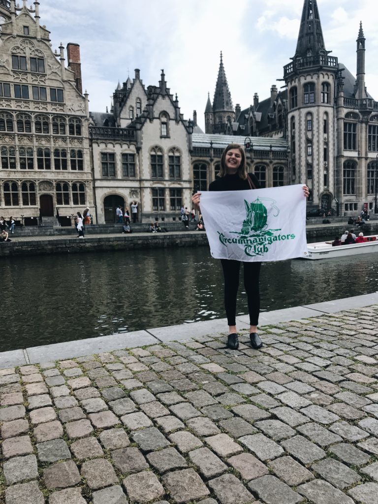 Fiona standing on a street in Belgium, holding up a flag next to a river.