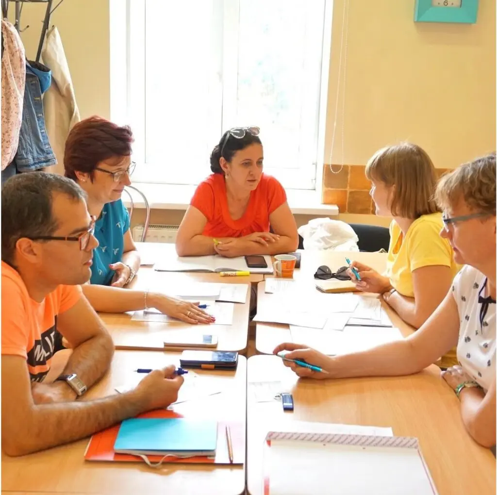 Alexander Begej in black-rimmed glasses, wearing orange t-shirt, sitting at long wooden table with others during Russian Immersion program.