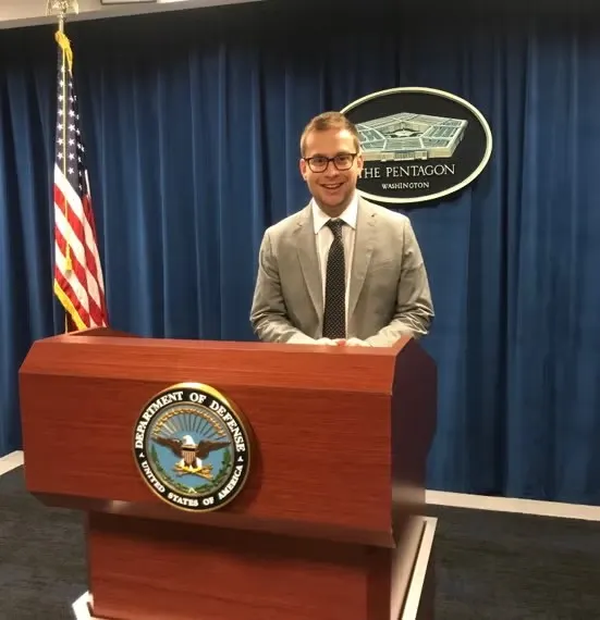 Ari Weisfuse standing at Pentagon podium, wearing gray suit with white shirt and navy blue tie.