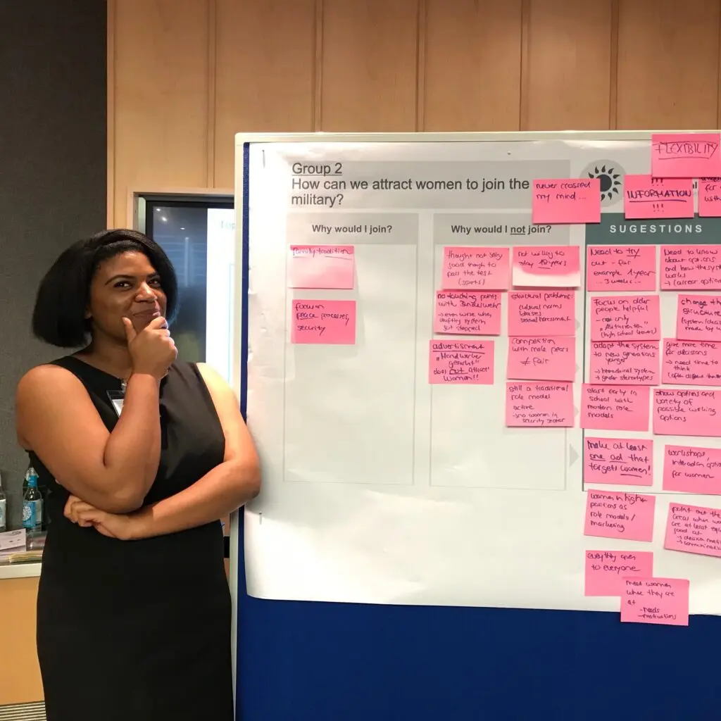 Meg McGee standing in black shift dress in front of presentation board at National Security conference in Germany.