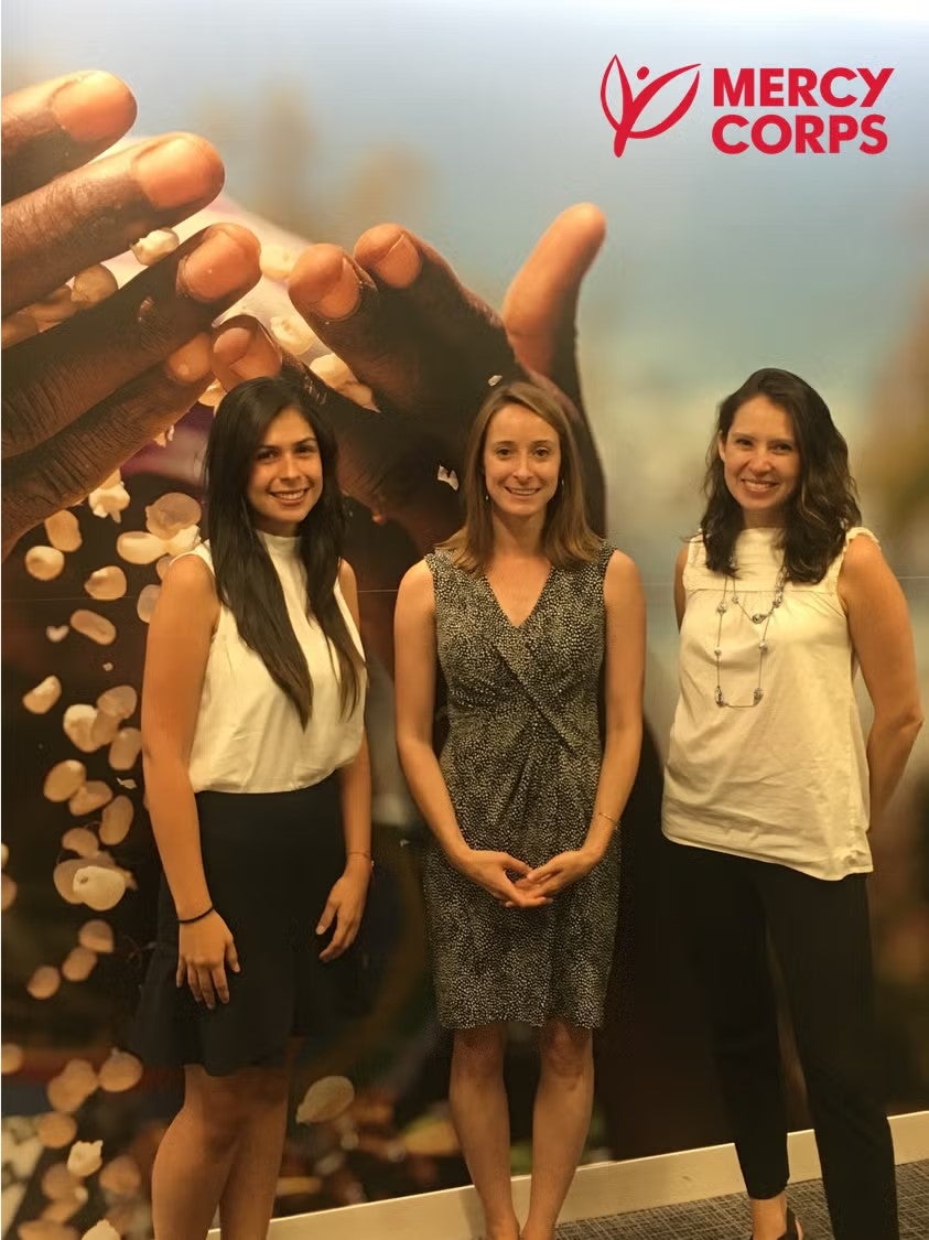 Silvia Rodriguez in white, sleeveless blouse and black pencil skirt, standing with Mercy Corps supervisors in front of poster representing Mercy Corps.