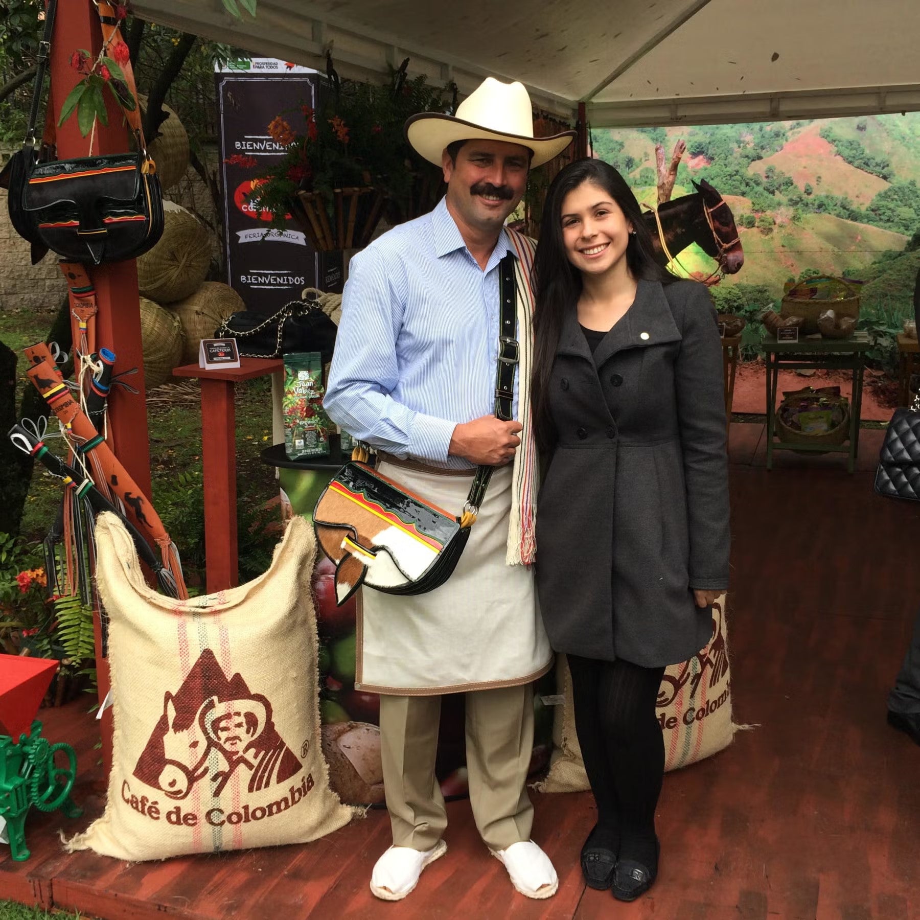 Silvia Rodriguez in Bogota, wearing black, wool coat and black tights, with black flats, standing next to farmer wearing ivory-colored cowboy hat, light blue button down shirt, khaki slacks, and white sneakers.