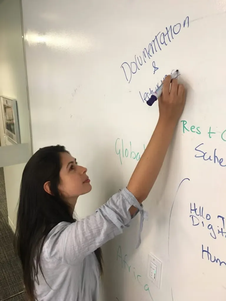 Silvia Rodriguez, wearing blue and white striped button down shirt, writing on whiteboard while working on project.
