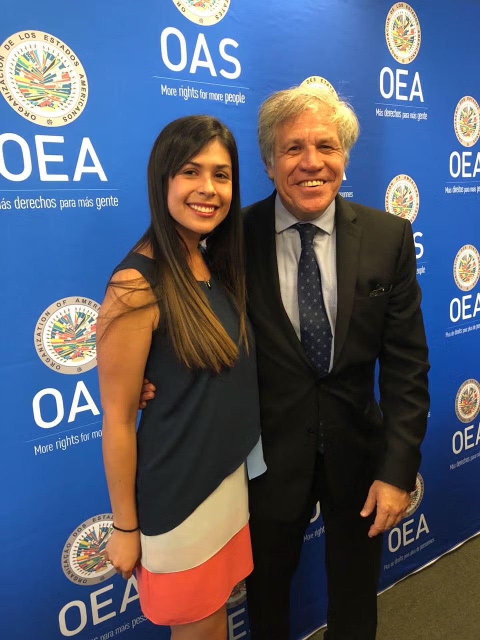 Silvia Rodriguez in blue, white, and orange shift dress, standing next to Luis Almagro, wearing a black suit, white button down shirt and navy blue tie.