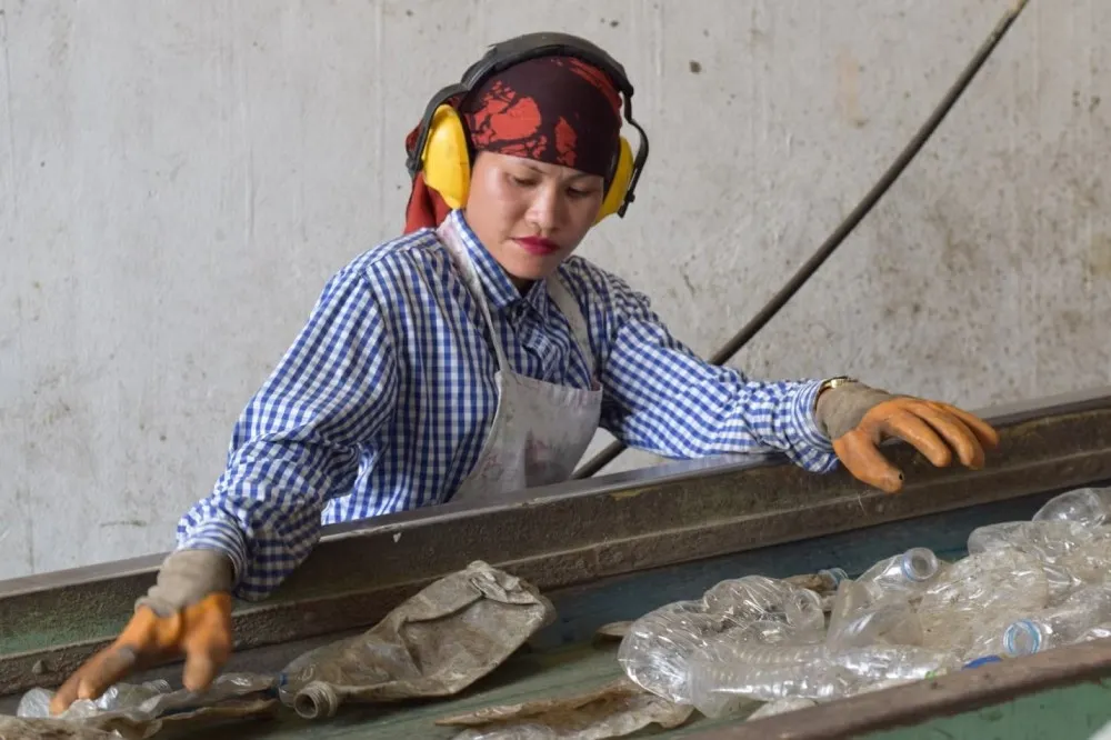 Young woman wearing earmuffs, a red headscarf, a blue checkered shirt, yellow work gloves, and an apron helping to move empty plastic bottles on a conveyor belt.