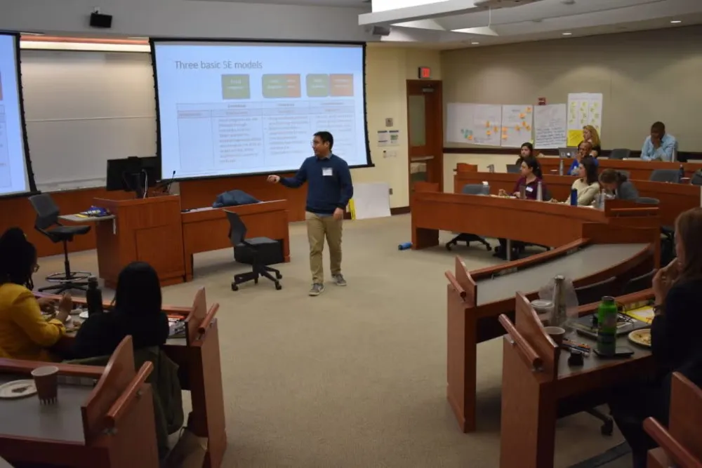 Young man in dark blue shirt and khaki chinos stands in front of classroom giving lecture to students during Social Innovation Lab competition.