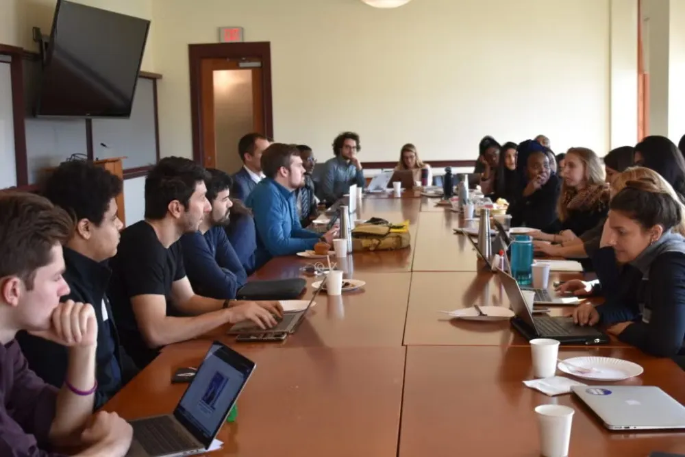 Many tens of students sitting around a long, brown table giving each other feedback on their projects for the Social Innovation Lab.