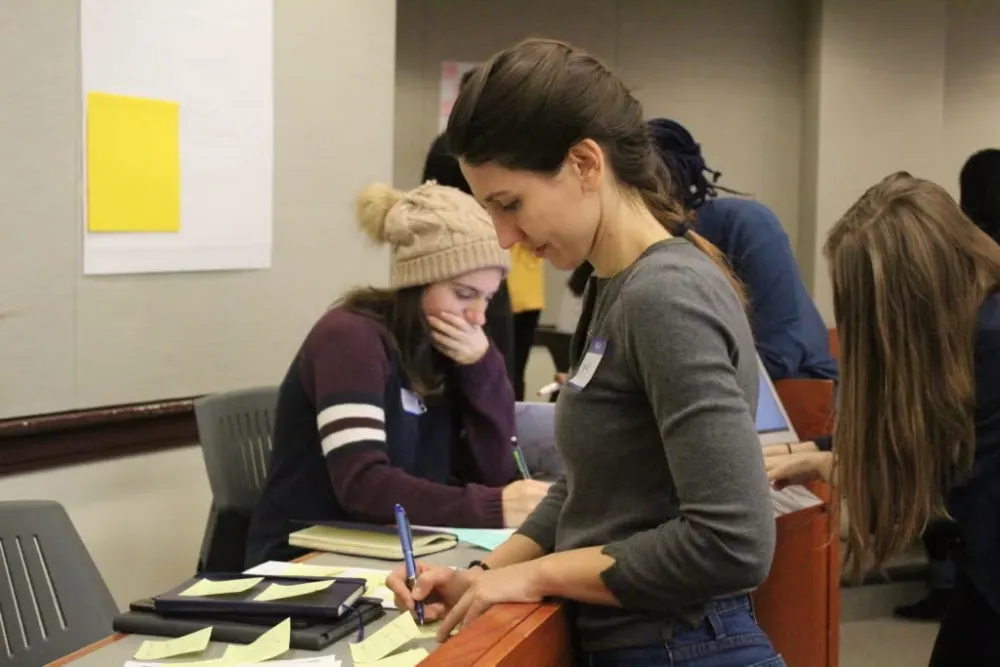 A young woman in a charcoal-colored long-sleeve shirt and jeans is writing on a yellow sticky note while working on her Social Innovation Lab competition project.