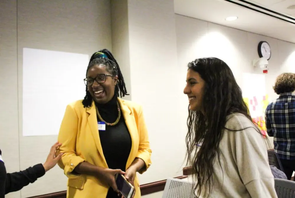 Two young women talking during Social Innovation Lap competition, one is wearing a yellow blazer and dark turtleneck; the other is wearing a cream-colored sweater.