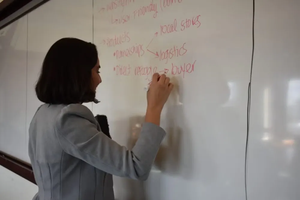 Young woman with dark bob, wearing gray blazer, standing at whiteboard, writing in red ink.