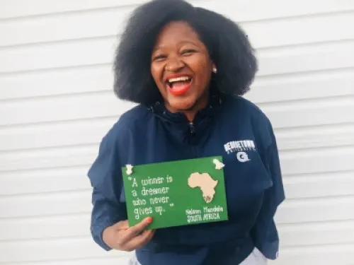 Maputi Botlhole with a big smile, standing in a navy blue Georgetown University jacket, holding a green sign with a quote from Nelson Mandela.