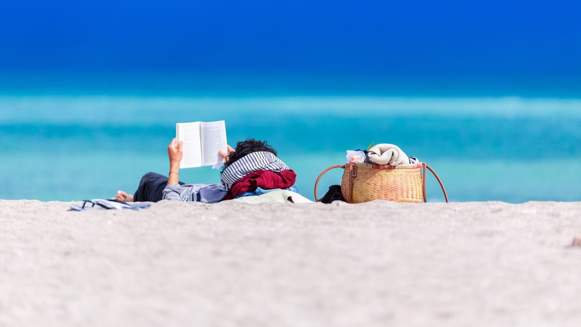 Person with dark hair in blue shirt, lying on sand reading book with ocean in the background.