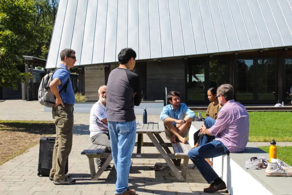 Five men and one woman dressed casually, sitting and standing around picnic table chatting.
