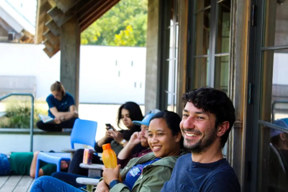 A group of young people are sitting in rocking chairs on a front porch. A young man with dark hair and a beard in a navy blue t-shirt and a young woman with dark hair in an olive green jacket, holding a small bottle of orange juice, smiling looking on at something in front of them. Others are sitting and relaxing, reading.