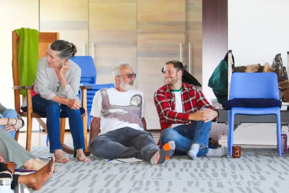 People casually sitting indoors talking at Ethics retreat. One man, in red flannel shirt and blue jeans, sitting on floor, talking with another man in white t-shirt and jeans. One woman in gray top and jeans, sitting in chair speaking with someone next to her, out of the photo's frame.
