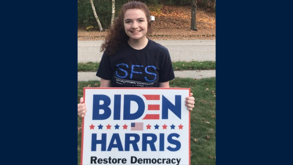 D'Amico poses in a front yard wearing an SFS t-shirt and holding a Biden-Harris yard sign.