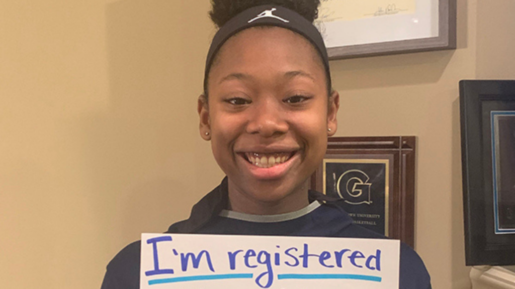 Cureton poses in her Hoyas Women's Basketball training kit with a handwritten sign reading "I'm registered".