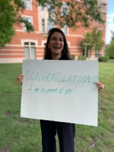 Prof. Grimm Arsenault pictured on campus with a sign saying "CONGRATULATION I am so proud of you."