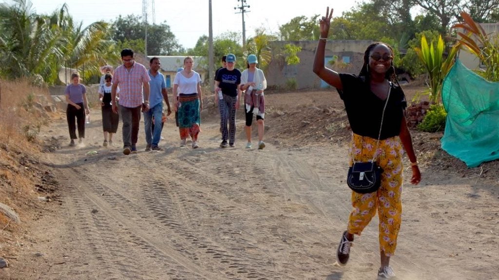 Travaly is walking through a residential area in Maharashtra, India. She is making the peace sign. Her classmates and professors walk behind her. 