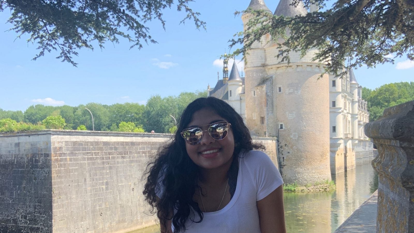 Menon is posing in front of a French castle. She is wearing mirrored sunglasses, a white shirt and a patterned skirt.