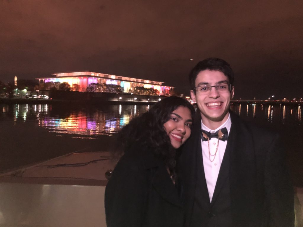 Menon and Felipe Lobo Koerich (SFS'21) are wearing formal clothes and posing on a boat in front of the Kennedy Center, which is lit up in rainbow lights