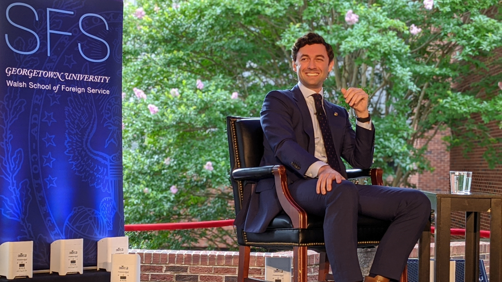 Jon Ossoff is seated on a stage in front of an SFS banner. He is looking toward the audience and smiling.