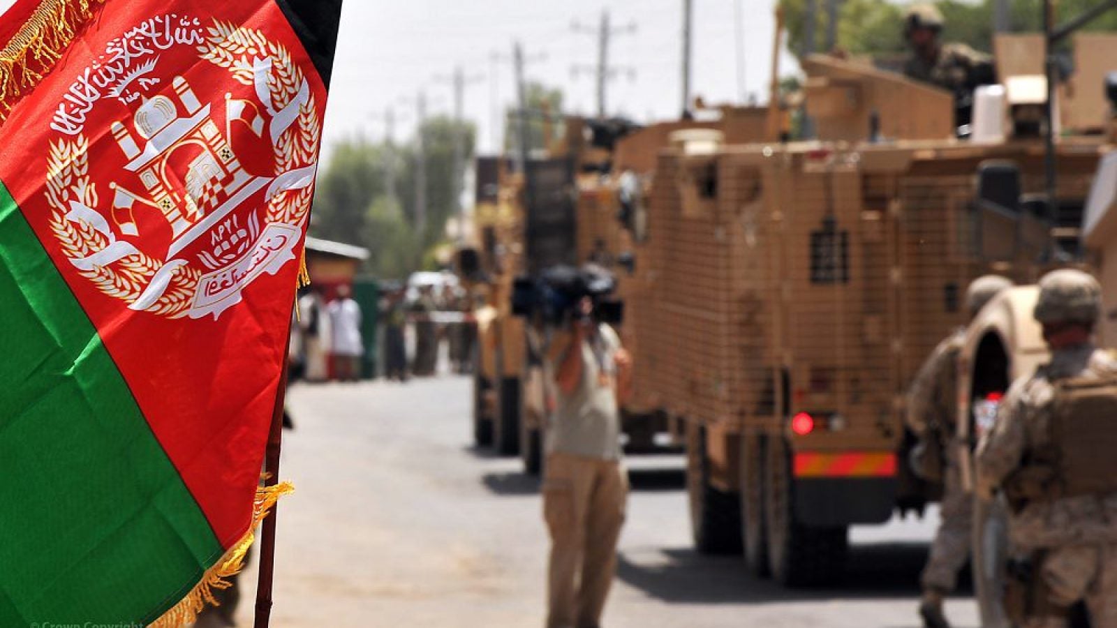 Afghan flag on the foreground, military troops and vehicles are blurred in the backgroud.