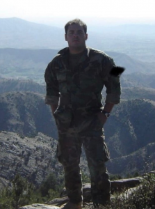 Torres, wearing an army combat uniform, standing in front of rolling hills in Afghanistan.