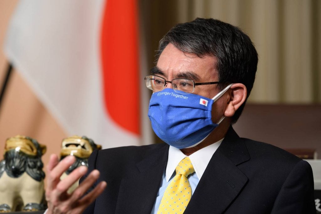 Kono wearing a blue face mask in front of Japanese flag.