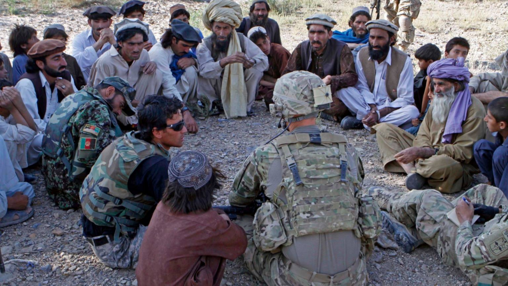 A U.S. Army officer talks with a group of Afghan men, sitting in a circle on the ground. An Afghan military interpreter is seated in the group.