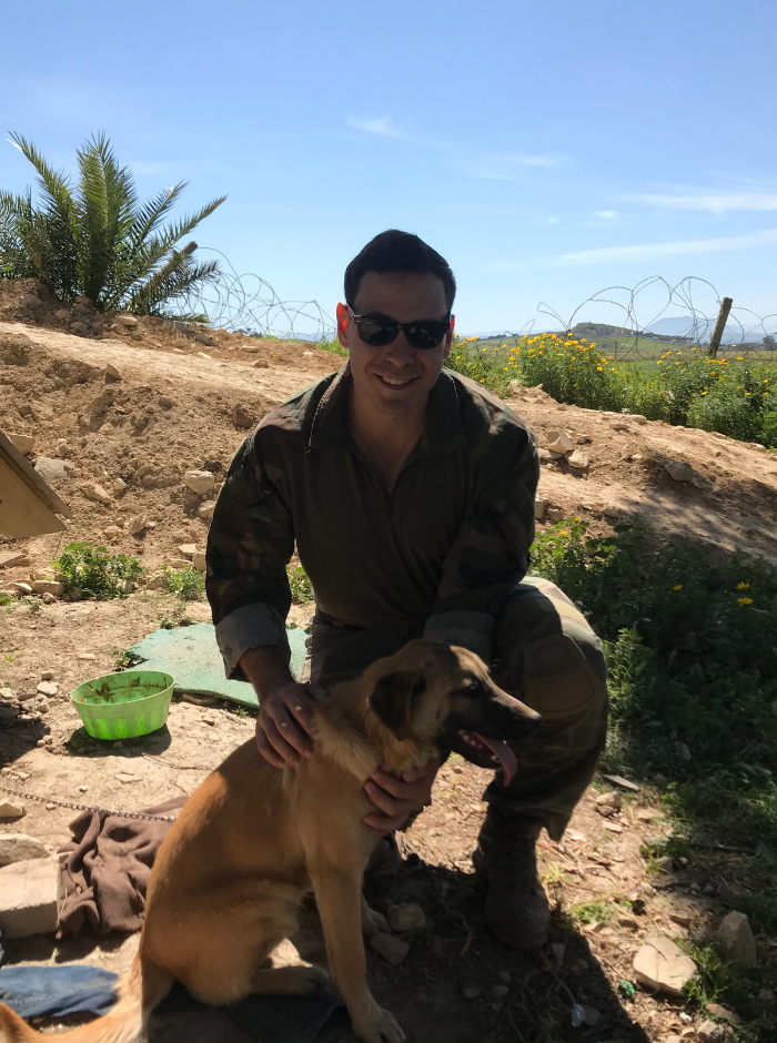 Zach Pyle wears combat gear. He is in a sandy landscape, petting a dog.