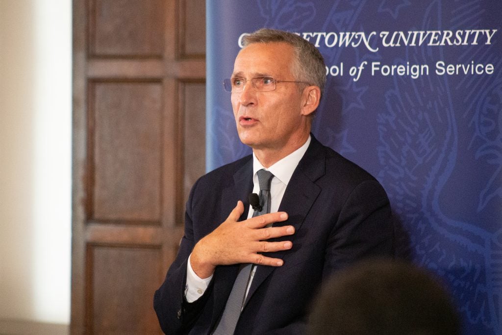 Stoltenberg sits in front of an SFS banner. He is speaking and is wearing a lapel mic.