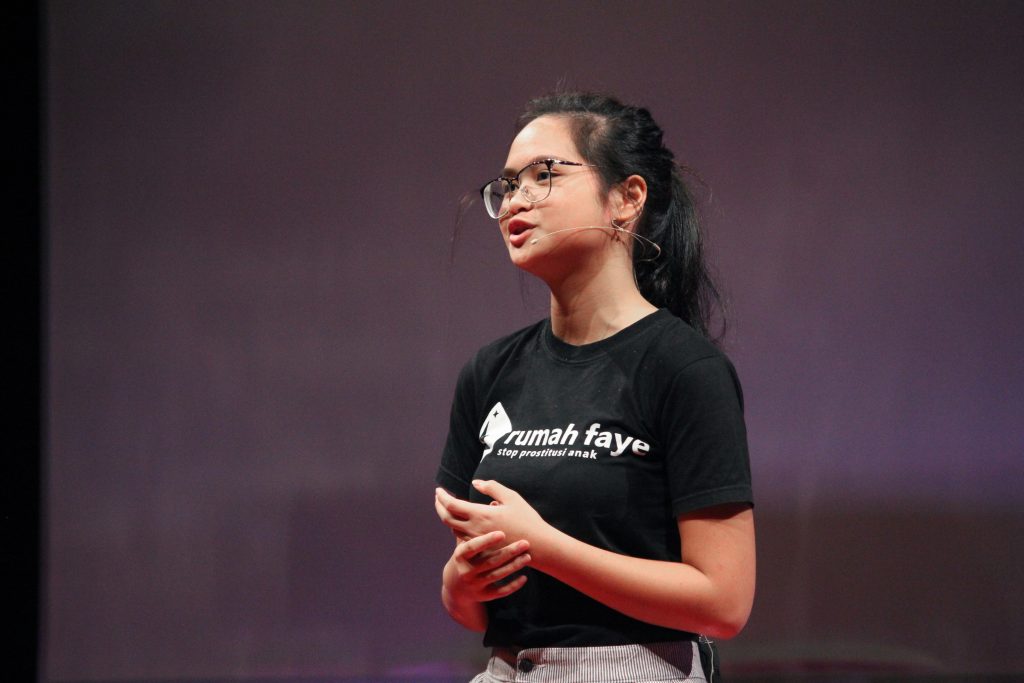 Faye Simanjuntak speaks on stage at a TED Talk event. She's wearing a black t-shirt with the name of her organization, Rumah Faye.