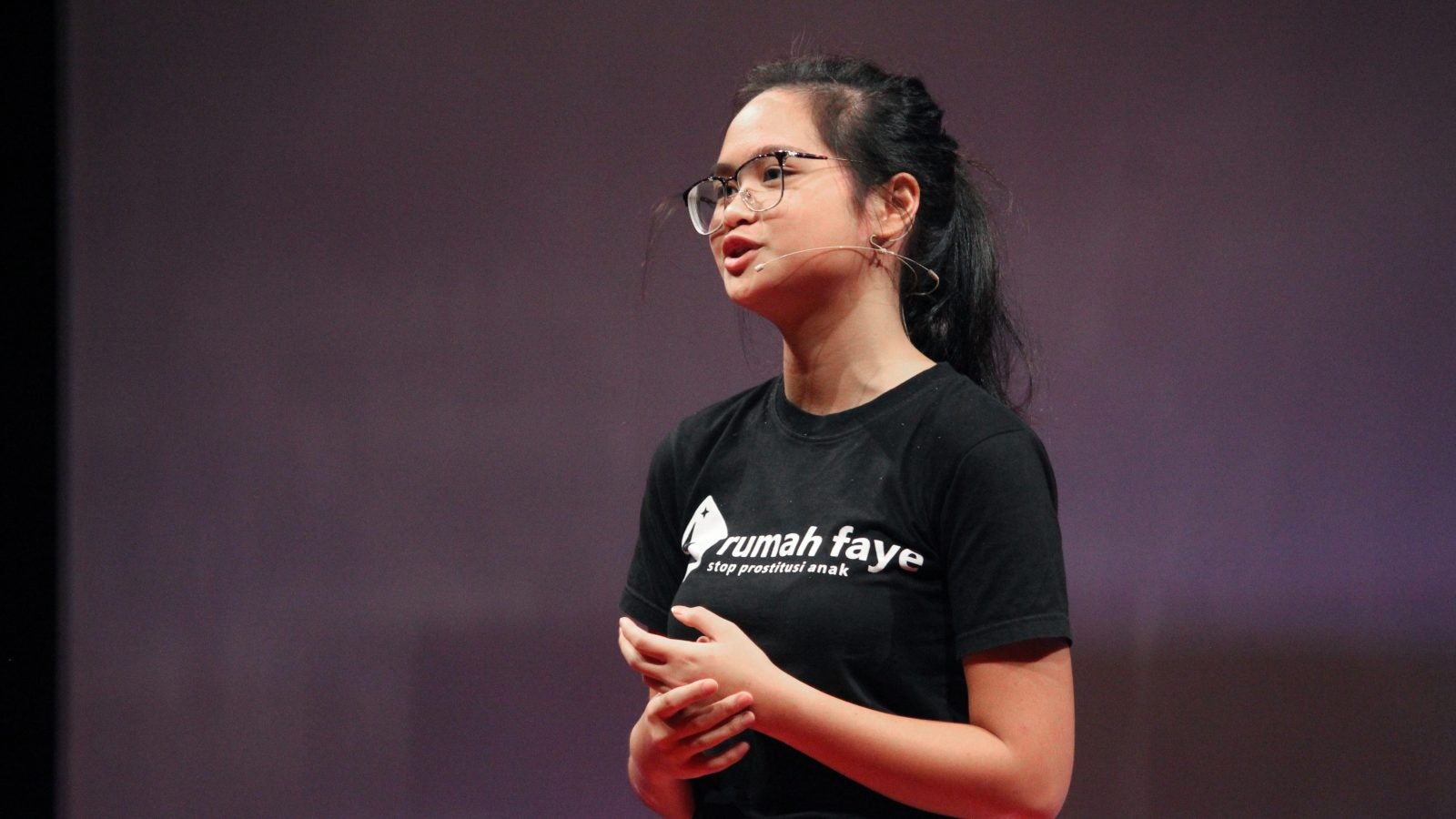 Faye Simanjuntak speaks on stage at a TED Talk event. She&#039;s wearing a black t-shirt with the name of her organization, Rumah Faye.