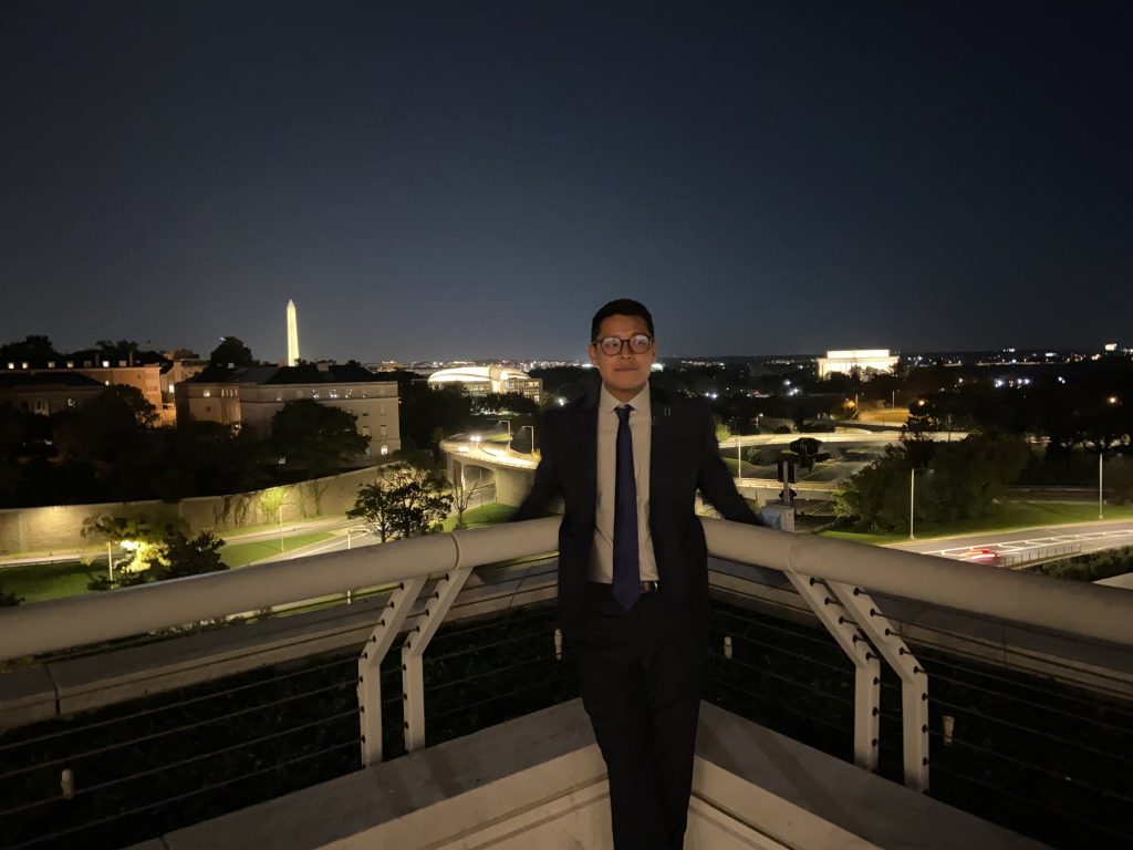 A photo of Sam in a suit, smiling on a rooftop with a view of the Washington Monument.