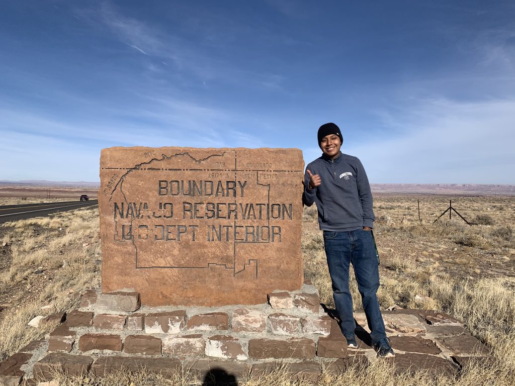 A photo of Sam wearing a Georgetown sweatshirt and smiling next to a sign for the Navajo Reservation