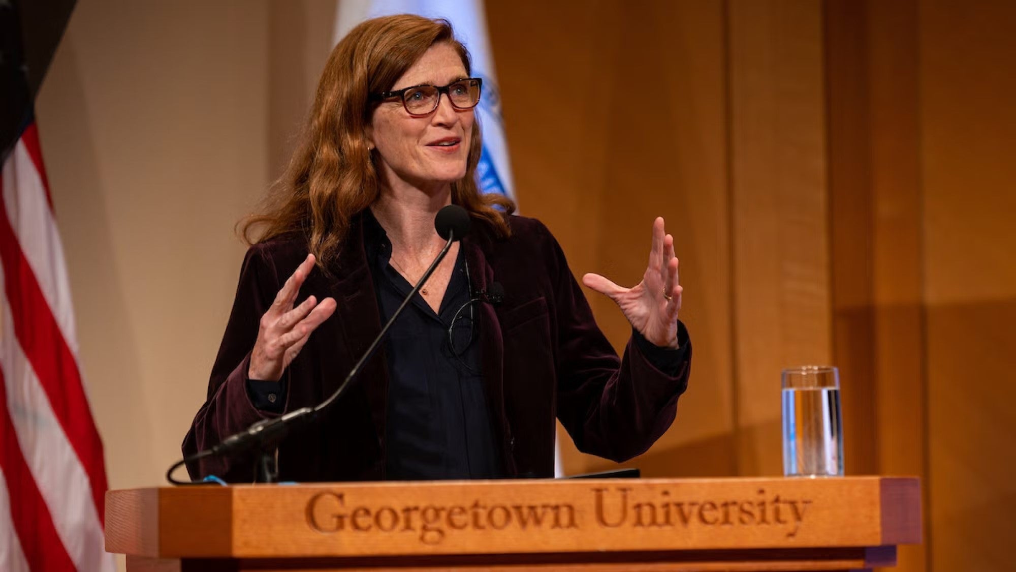Samantha Power in black suit standing at podium, next to American flag, with light brown, wooden wall behind her. 