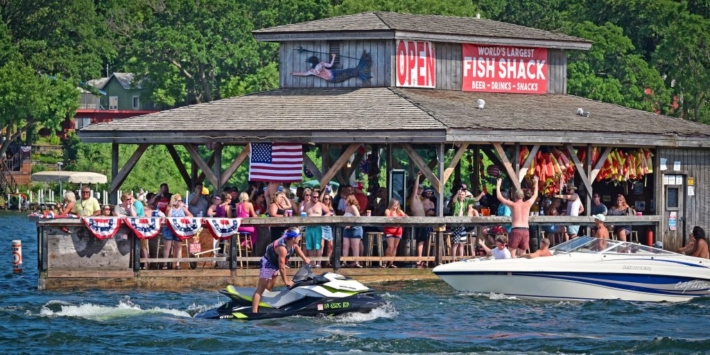 A photo of a crowded lakeside bar and restaurant during the summertime. People drive past in boats and jetskis.