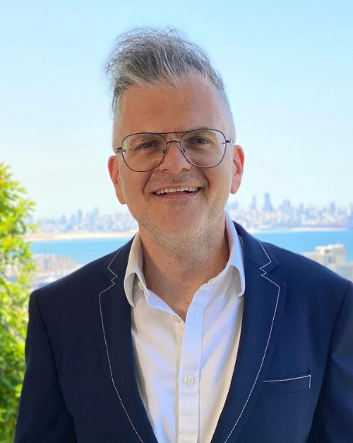 Photo of Deputy Chief of Mission Richard Michaels (SFS'96) smiling and wearing a suit against a sunny outdoor backdrop.