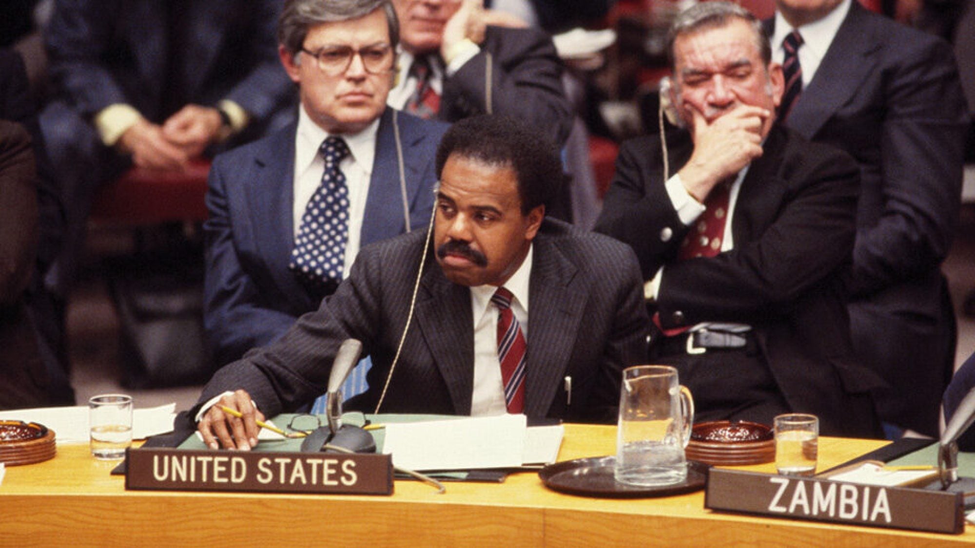 A photo of U.S. Ambassador to the UN Donald F. McHenry (PHD&#039;63) sitting behind a desk with a microphone and a &quot;United States&quot; placard on it.