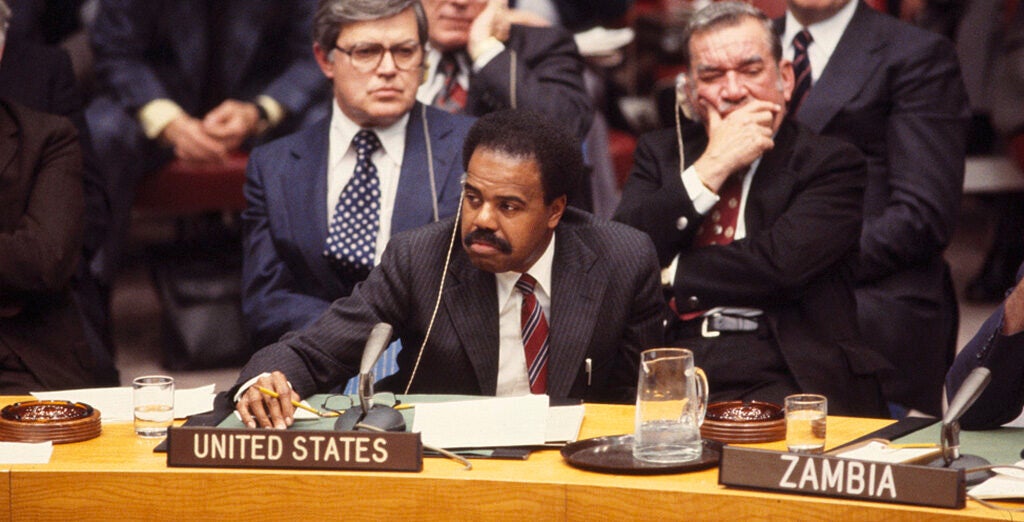 A photo of U.S. Ambassador to the UN Donald F. McHenry (PHD'63) sitting behind a desk with a microphone and a "United States" placard on it.