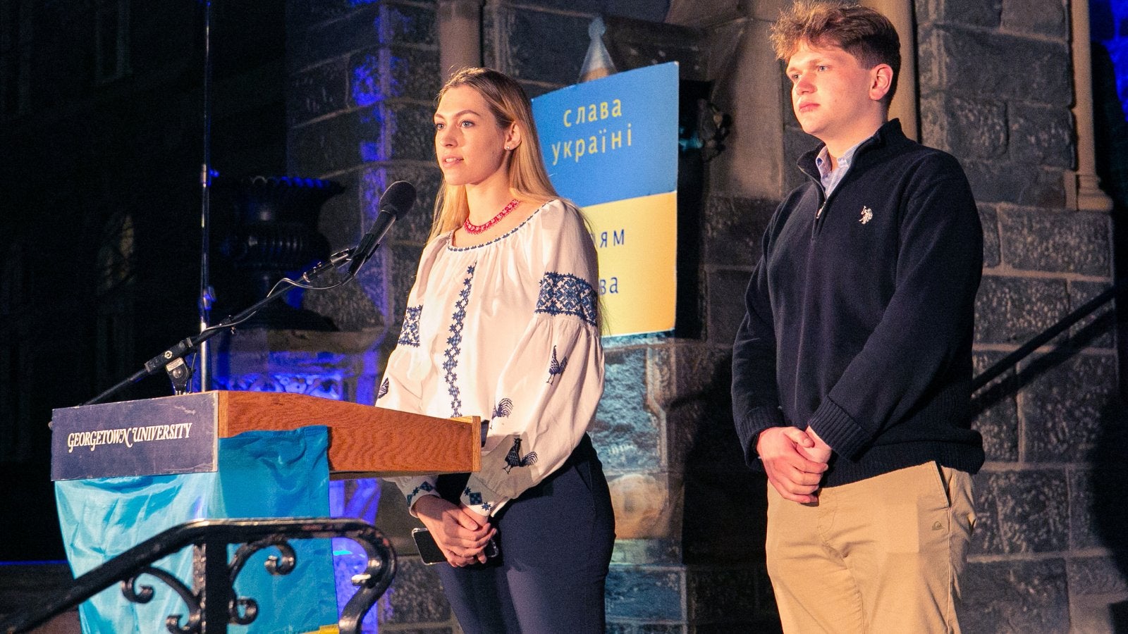 Two students stand behind a podium on the Healy Hall steps. A banner behind them features the colors of the Ukrainian flag.