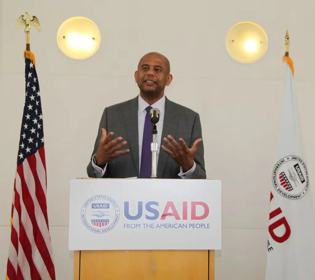 Gary Barrett in suit standing at United States Agency for International Development podium, with palms in front of his chest, facing up, giving a speech.