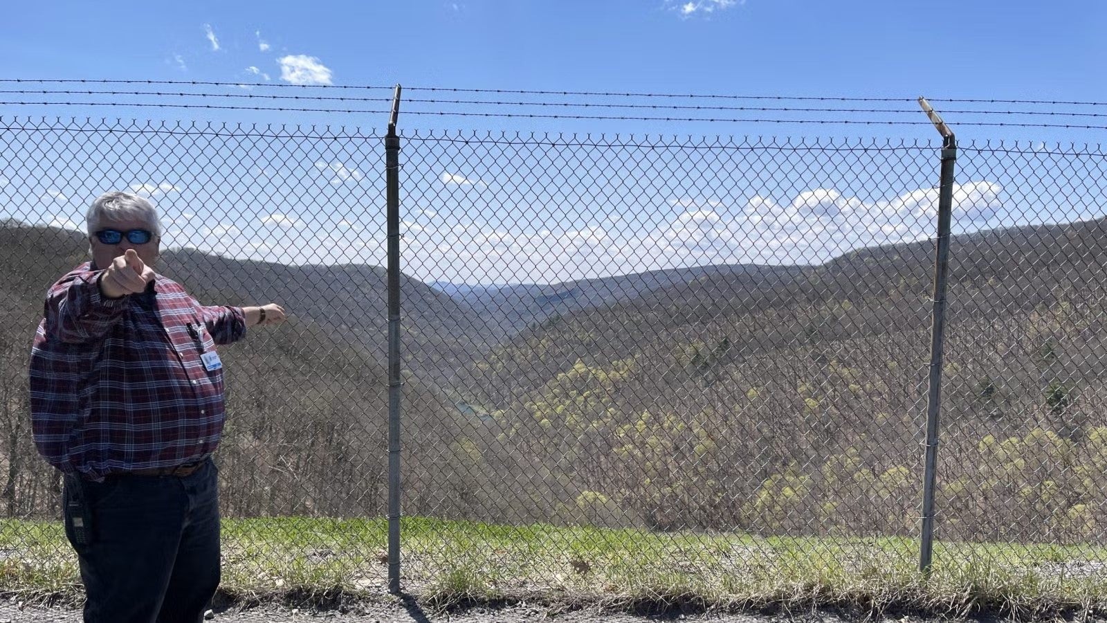 Man standing, holding on to fence, pointing towards camera, wearing a plaid button down shirt and dark blue jeans.