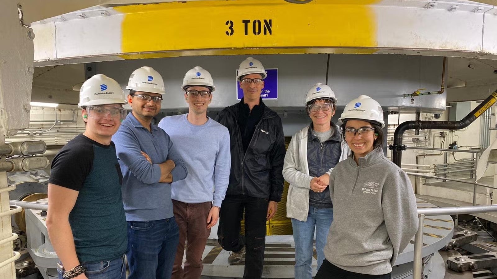 Five students in casualwear and white hardhats inside plant facility, standing together.